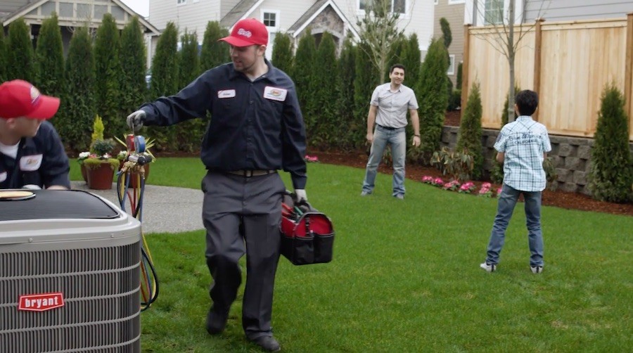 Two AC technicians service a Bryant unit while a parent and child play in background.