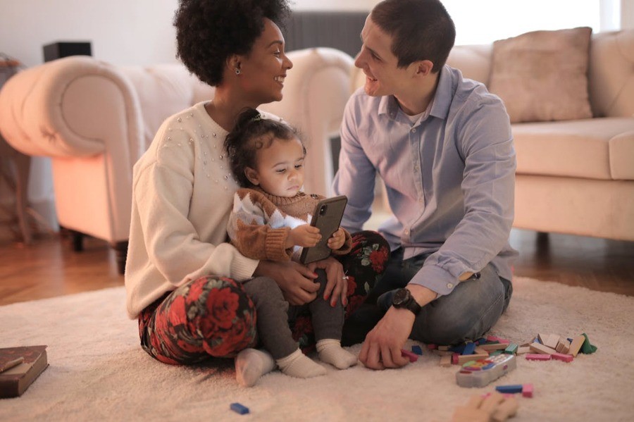 A couple sits on their living room rug playing with a baby who has toys scattered on the floor. 